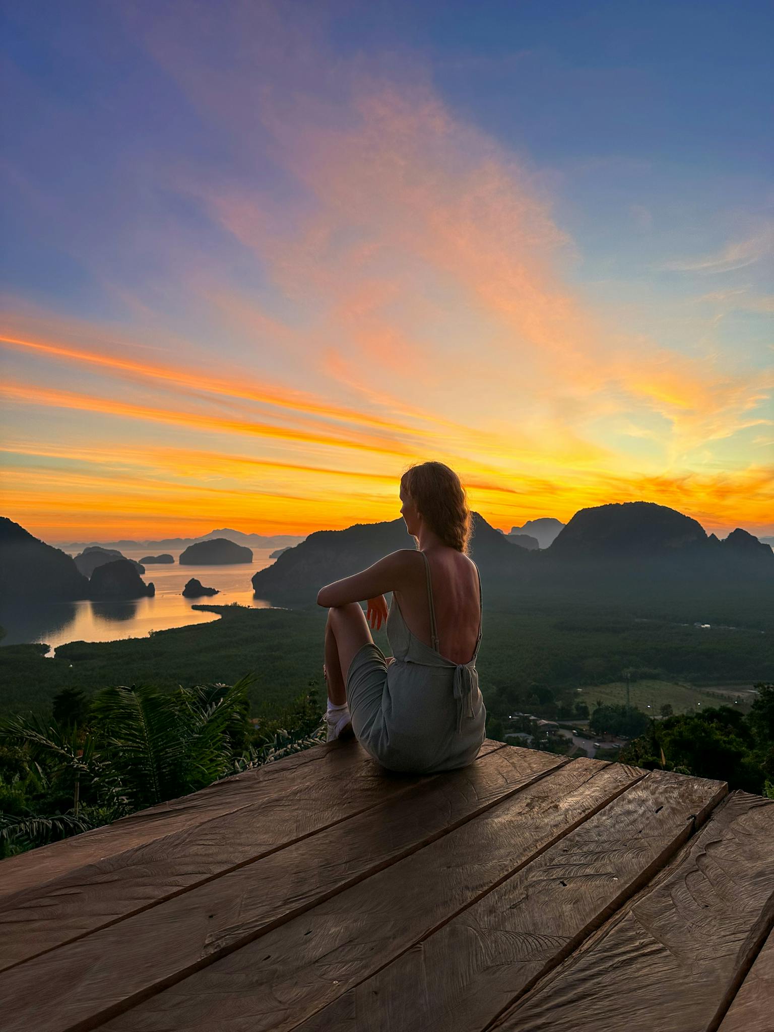 A woman sits on a platform enjoying a breathtaking sunrise over a scenic bay with limestone cliffs.
