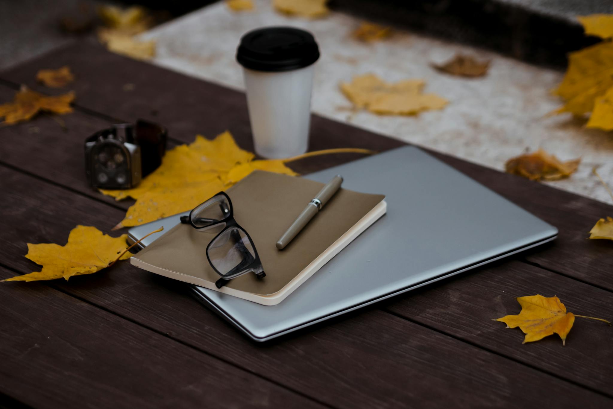 Outdoor autumn workspace setup with laptop, notebook, pen, and coffee on a wooden table with fallen leaves.
